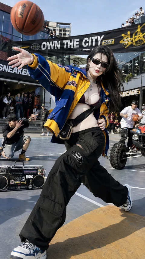 A person in sunglasses, a blue-and-yellow jacket, and loose black trousers reaches toward a basketball on a busy shopping street.